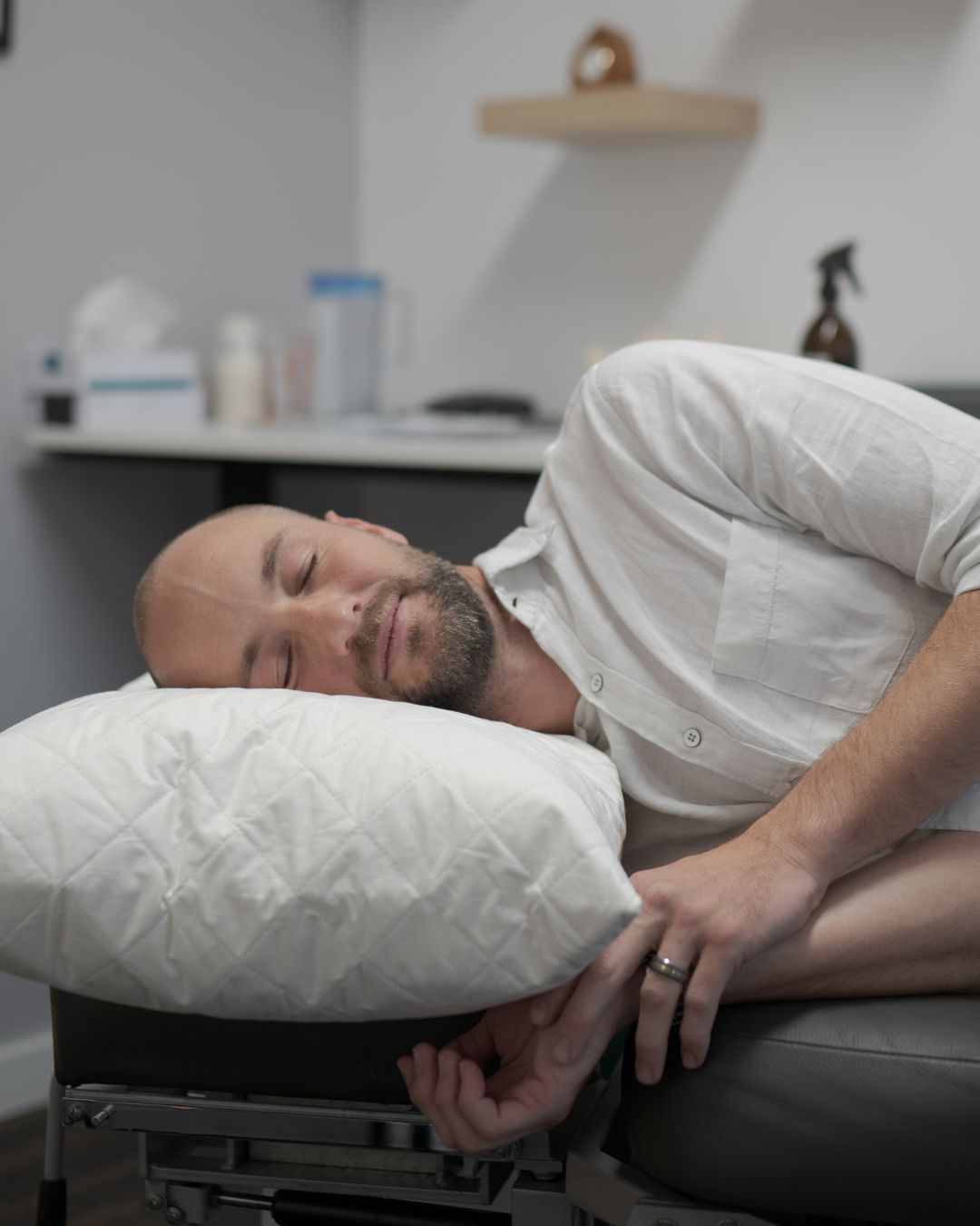Man lying on his side on a white pillow in a clinic room, resting with eyes closed.