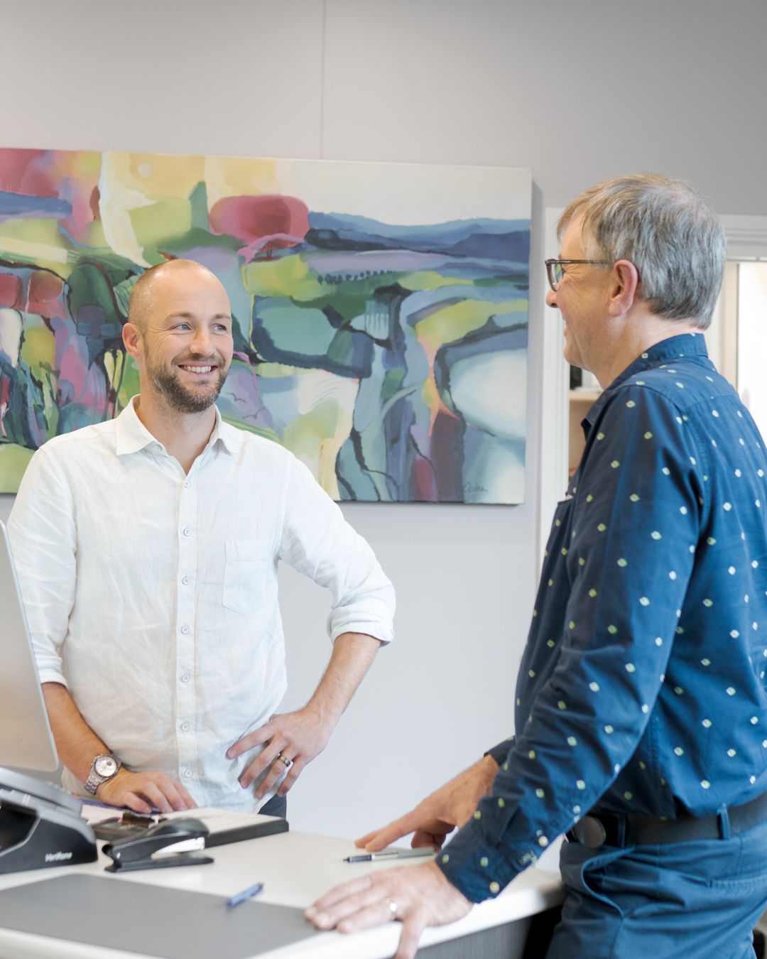 Two men talking across a desk in an office, with colorful abstract art on the wall.