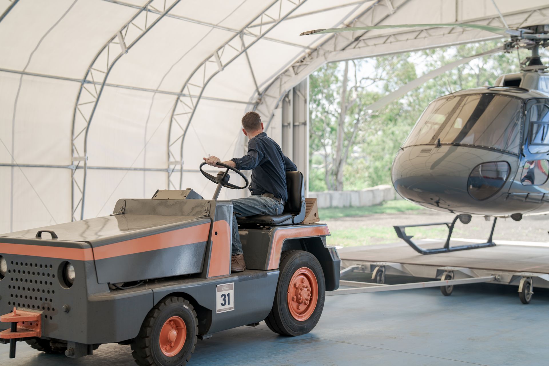 Person driving a vintage tractor beside a helicopter inside a hangar
