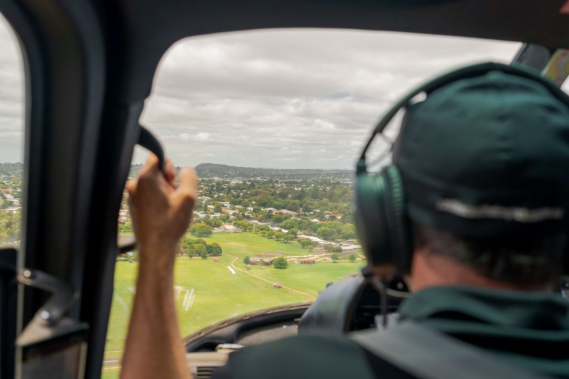 Inside a helicopter cockpit, a pilot looks out over a green landscape through the open door.