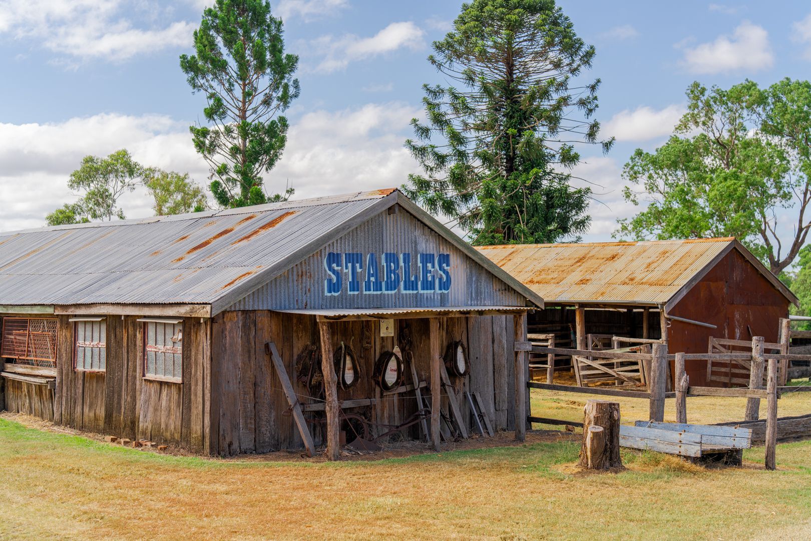Rustic wooden stables with a blue sign, weathered roof, and trees under a partly cloudy sky