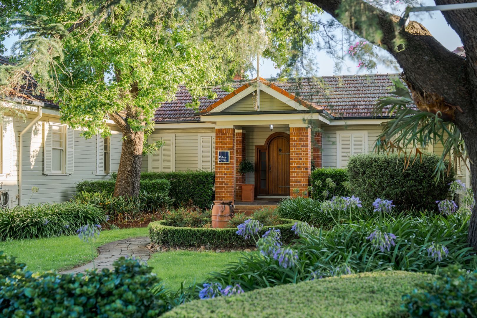Brick bungalow home with a front porch, white shutters, and a lush green garden under large trees