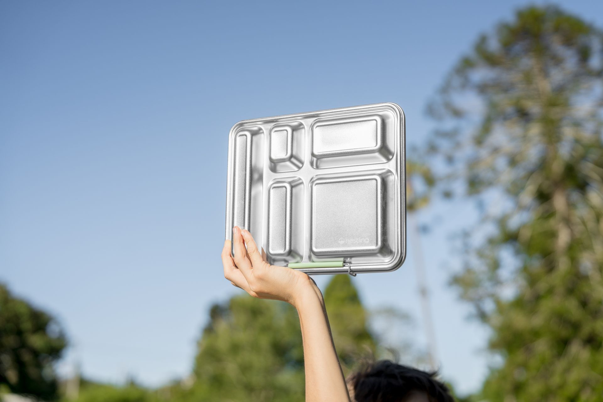 Hand holding a silver metal tray against a blue sky and green trees