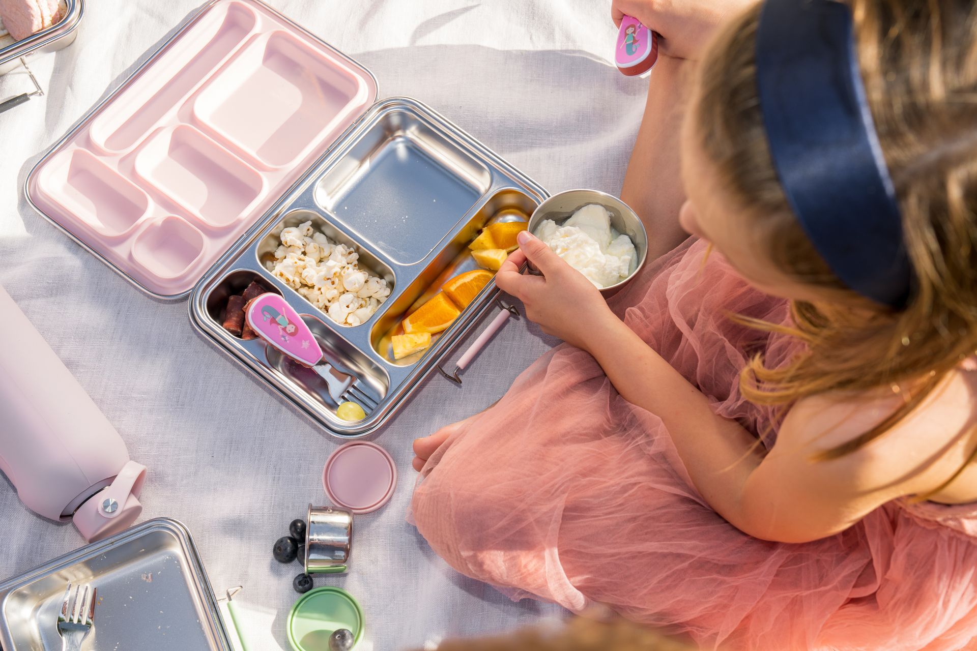 Child in a pink dress packing lunch from a colorful bento box on a pastel blanket