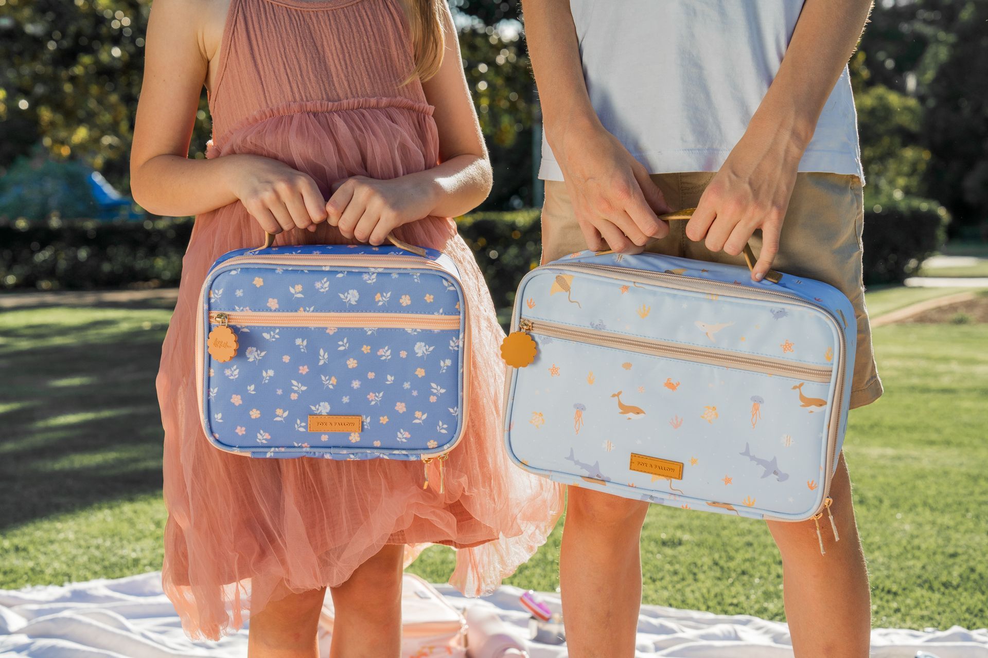 Two people holding pastel blue lunch bags with orange handles outdoors on grass.