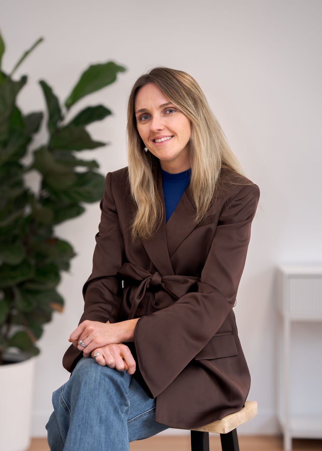 Blonde woman in a cream blazer and gray pants sitting on a stool, smiling against a white background