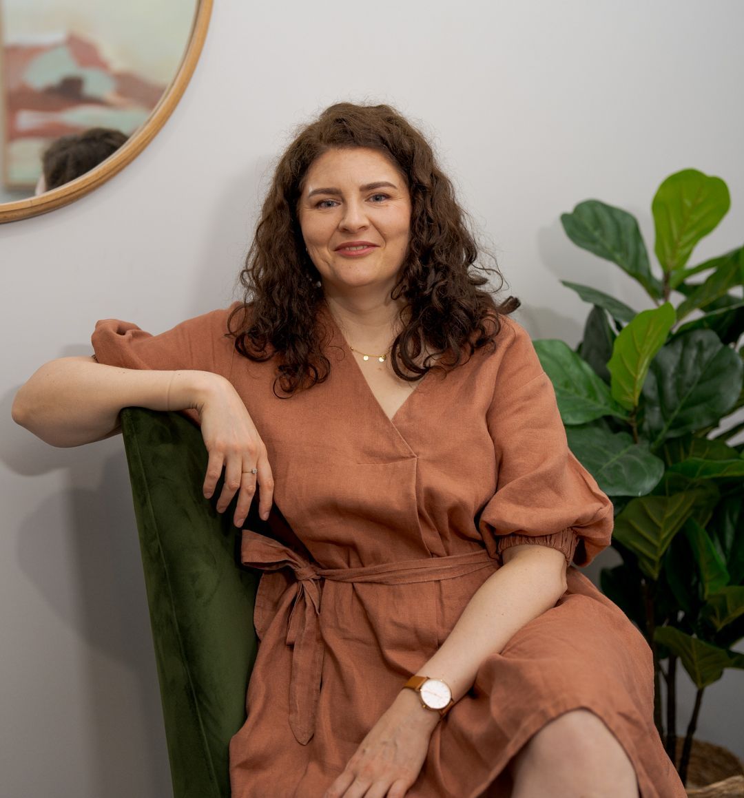 Woman in a rust-colored dress seated on a green chair beside a leafy plant, smiling in a bright room.