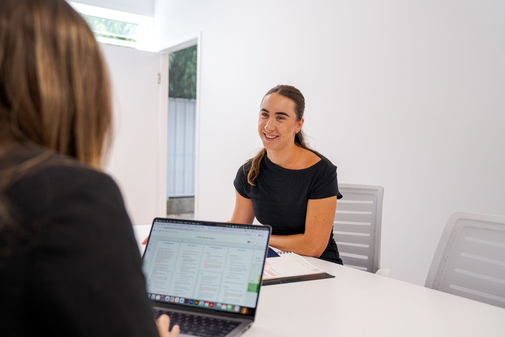 Two people in a bright office meeting, one smiling across a table with a laptop open.