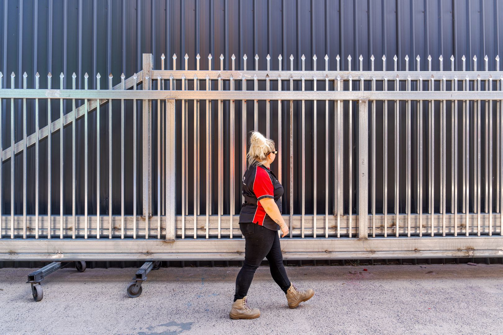 Person in red shirt walking beside a metal fence with wheeled barriers nearby.