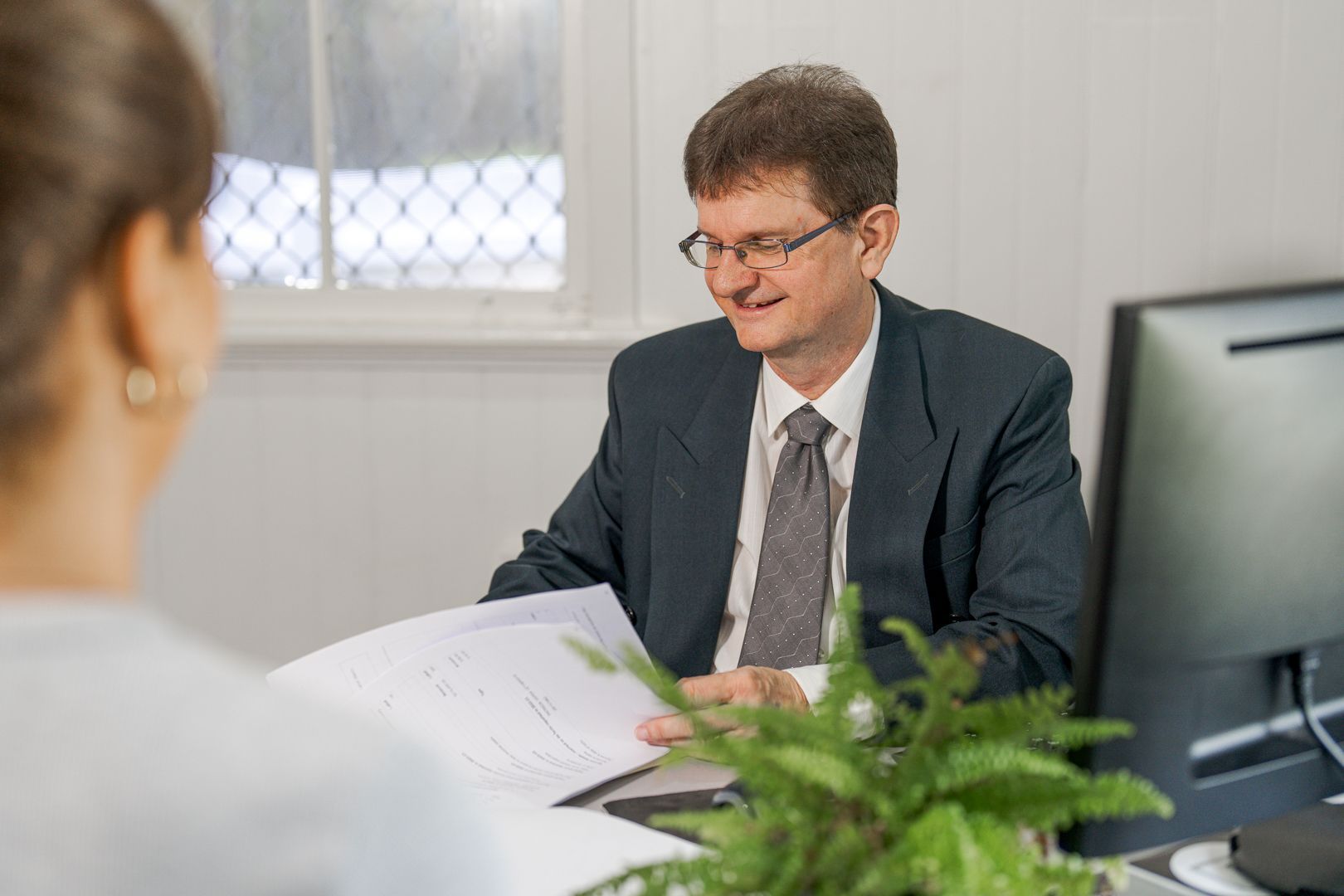 Office meeting with a man reviewing papers across a desk from another person, with a computer and plant nearby