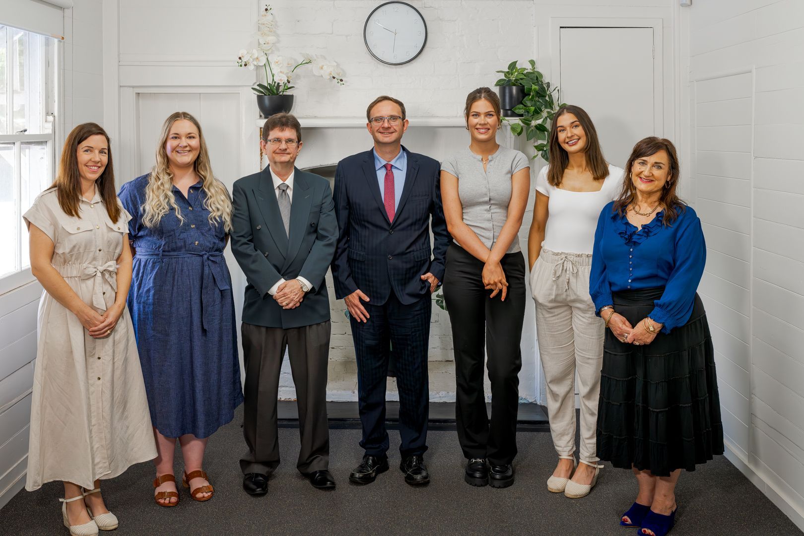 Seven people standing indoors in formal attire, posing together in front of a white wall and clock.
