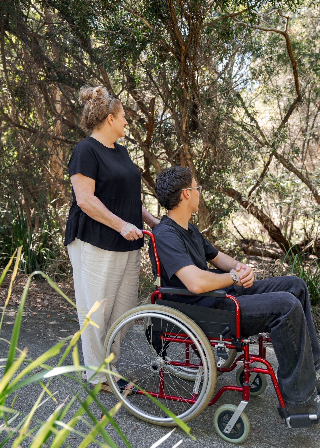 Two people outdoors, one standing beside a wheelchair with the seated person looking to the right.