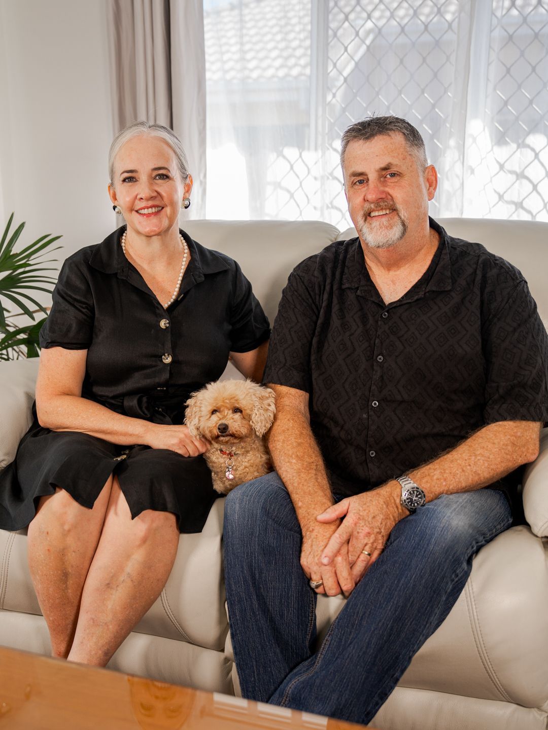 Two people sit on a white couch with a small tan dog in a bright living room.