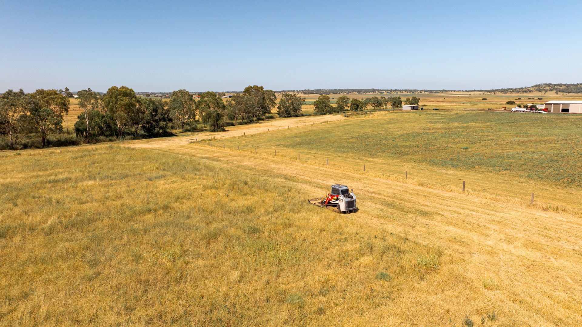 Small red tractor mowing a golden field under a clear sky, with trees along the horizon.