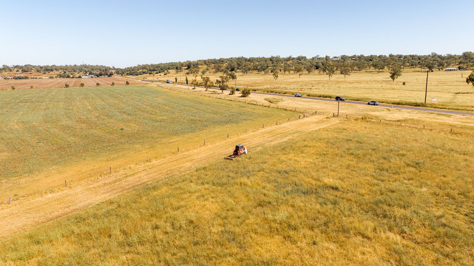 Dry grassy field with a dirt road, scattered trees, and a few small figures in the distance under a clear sky