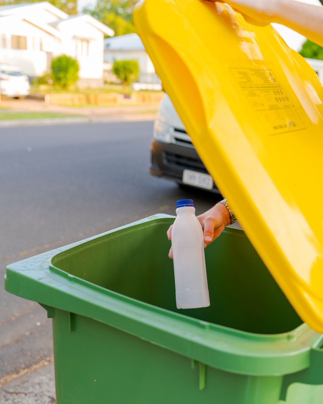 Hand placing a plastic bottle into a green recycling bin with a yellow lid on a street side