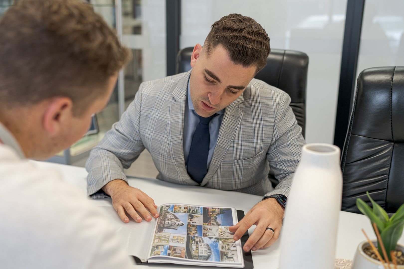 Two men in suits discuss printed materials at a desk in a modern office