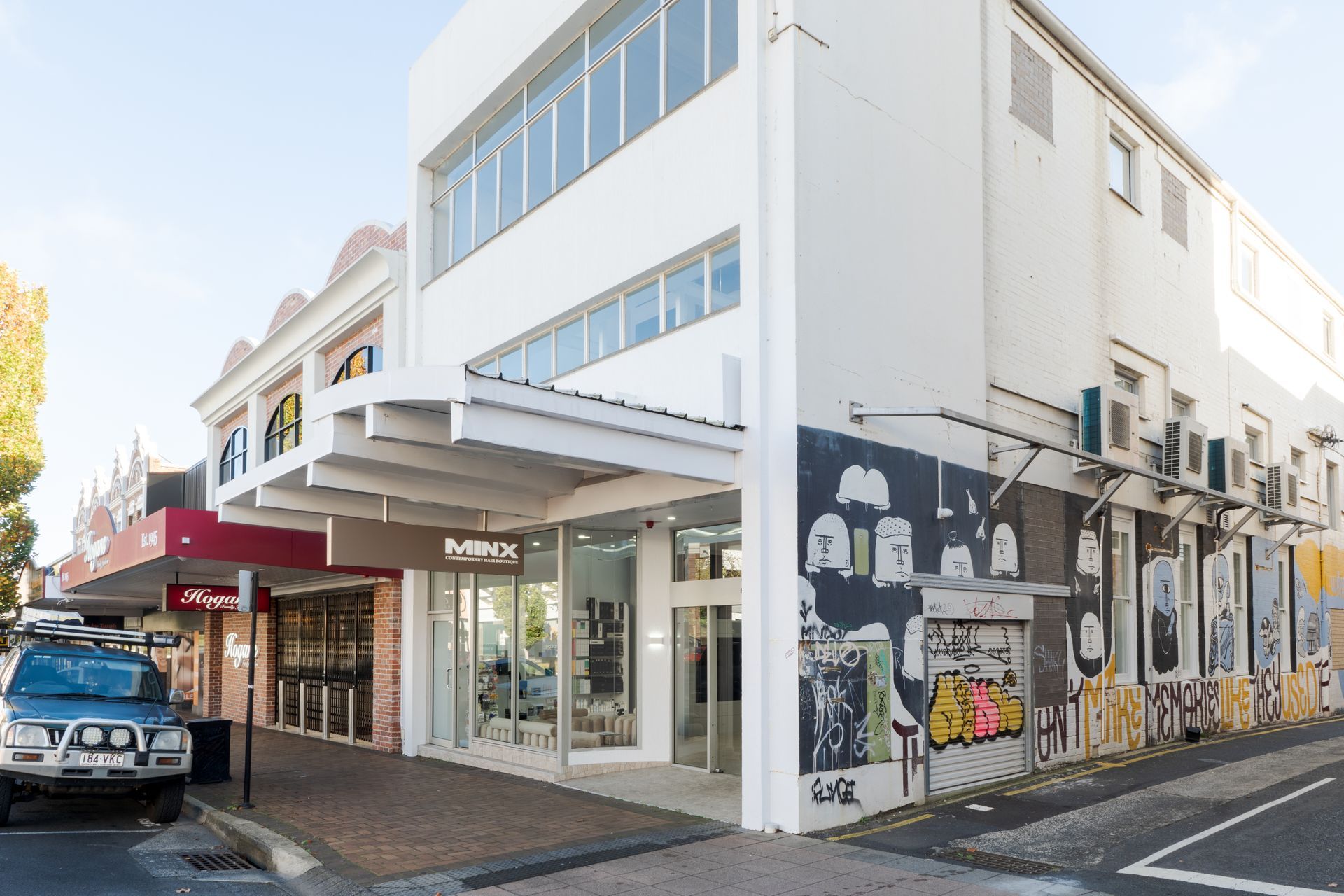 Street corner with modern white storefronts, murals on the wall, and a parked blue car.