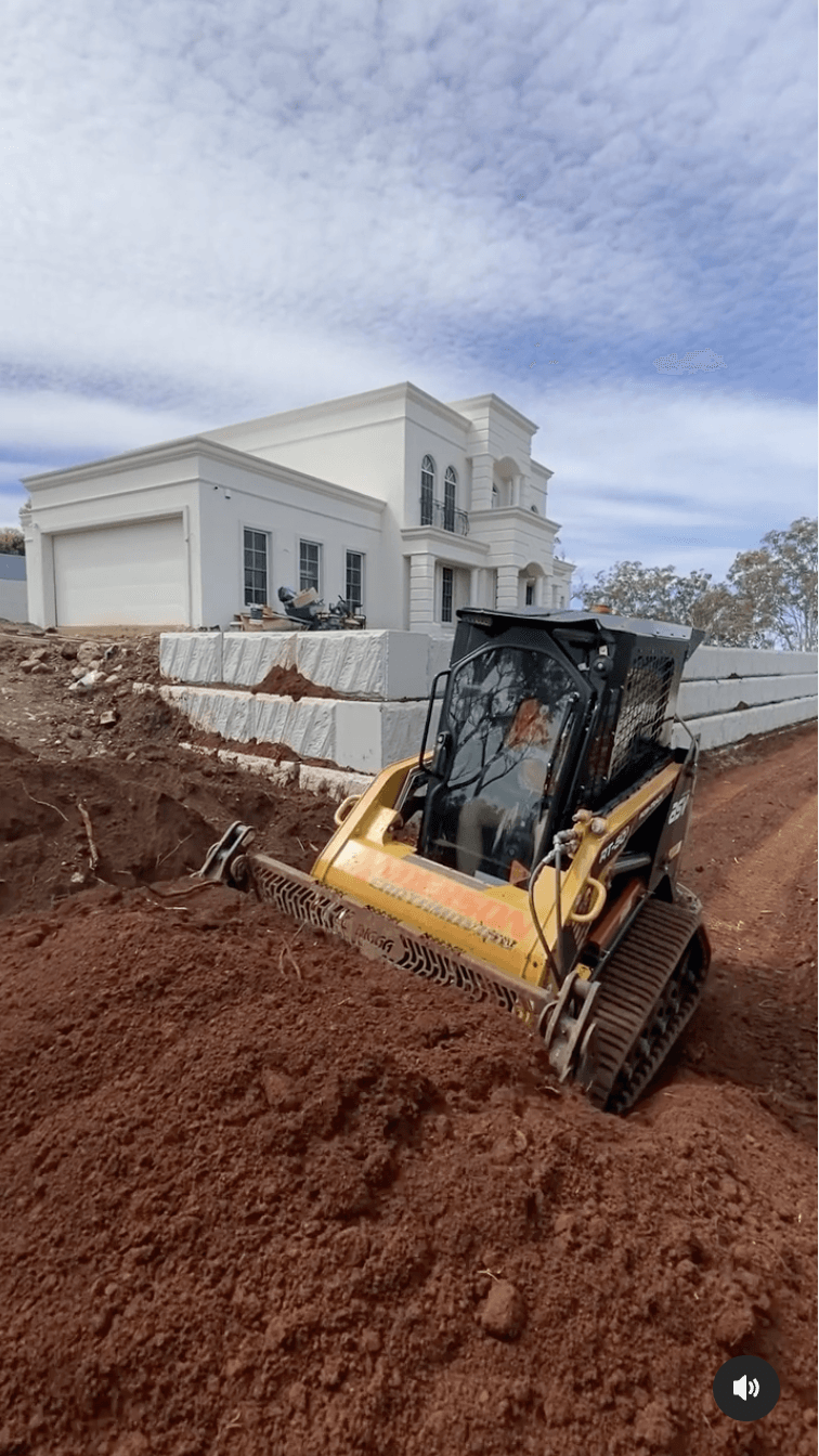 Yellow skid steer on red dirt beside a white house under construction with cloudy sky.