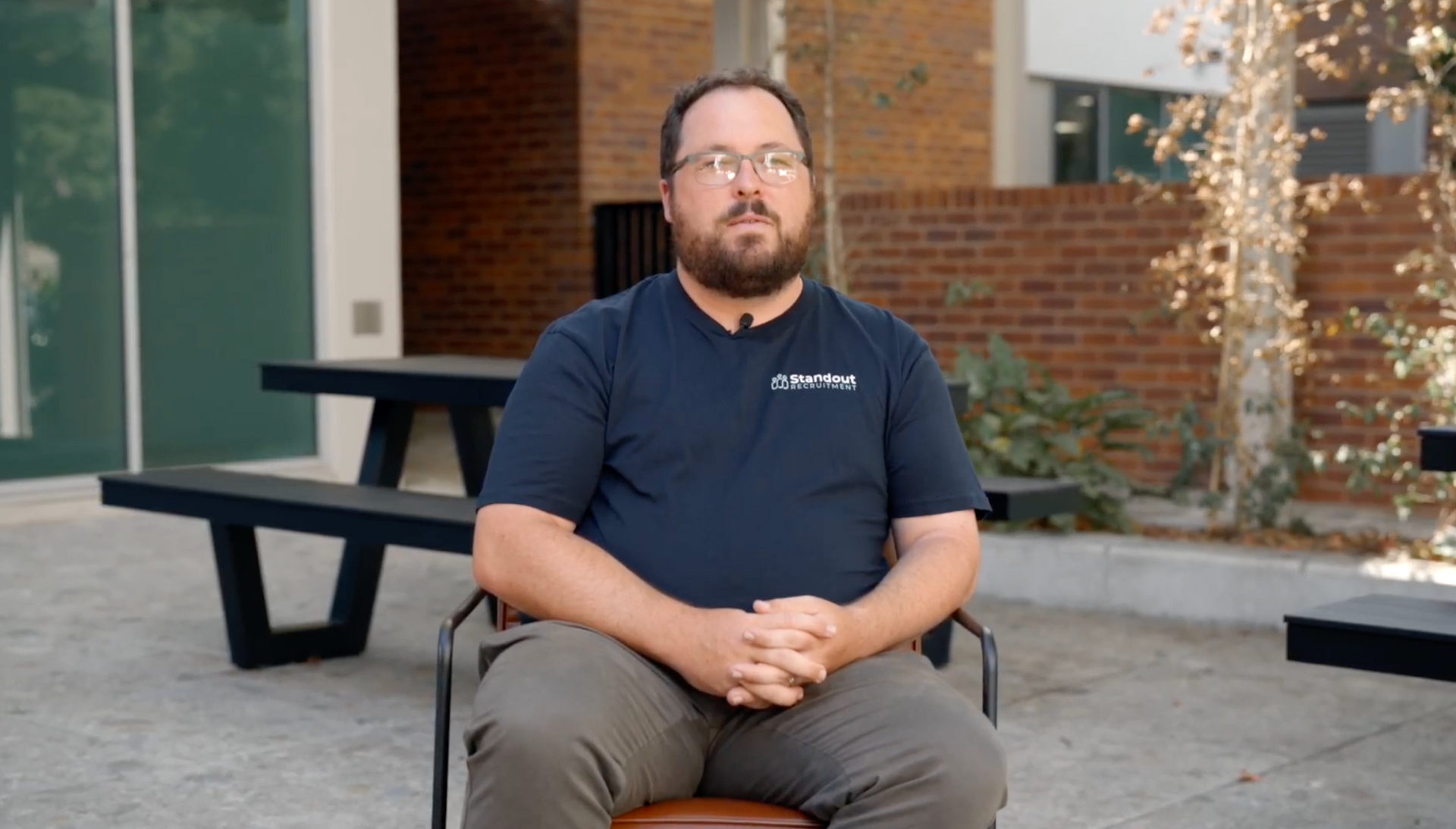 Man seated outdoors on a bench in front of a building, wearing a navy shirt and khaki pants.