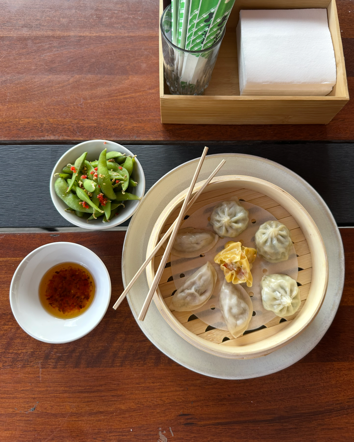 Steamed dumplings in a bamboo basket with chopsticks, dipping sauce, and a small side salad on a wooden table