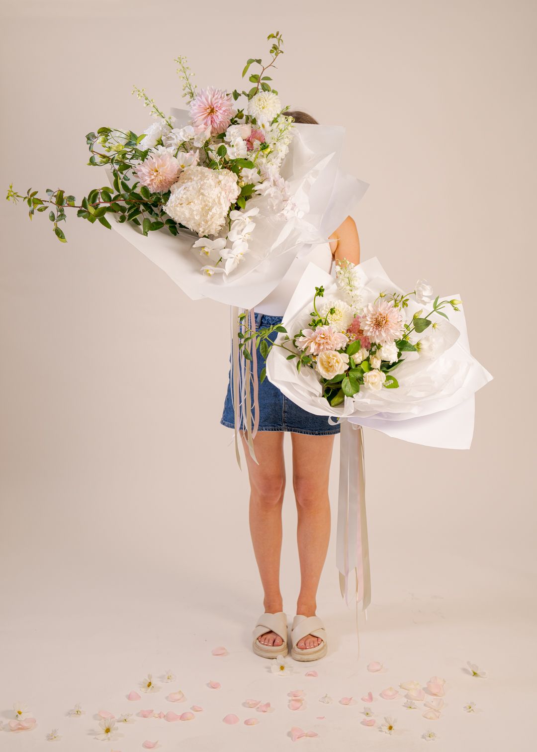 Person holding two large white flower bouquets against a beige backdrop, petals scattered on the floor