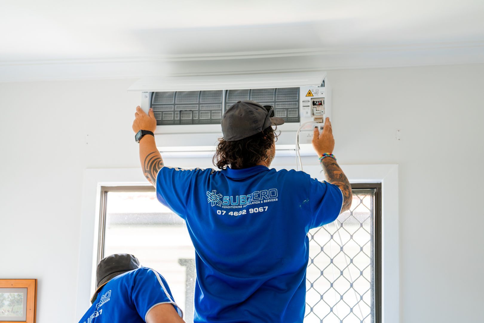Workers installing a wall-mounted air conditioner above a window in a white room