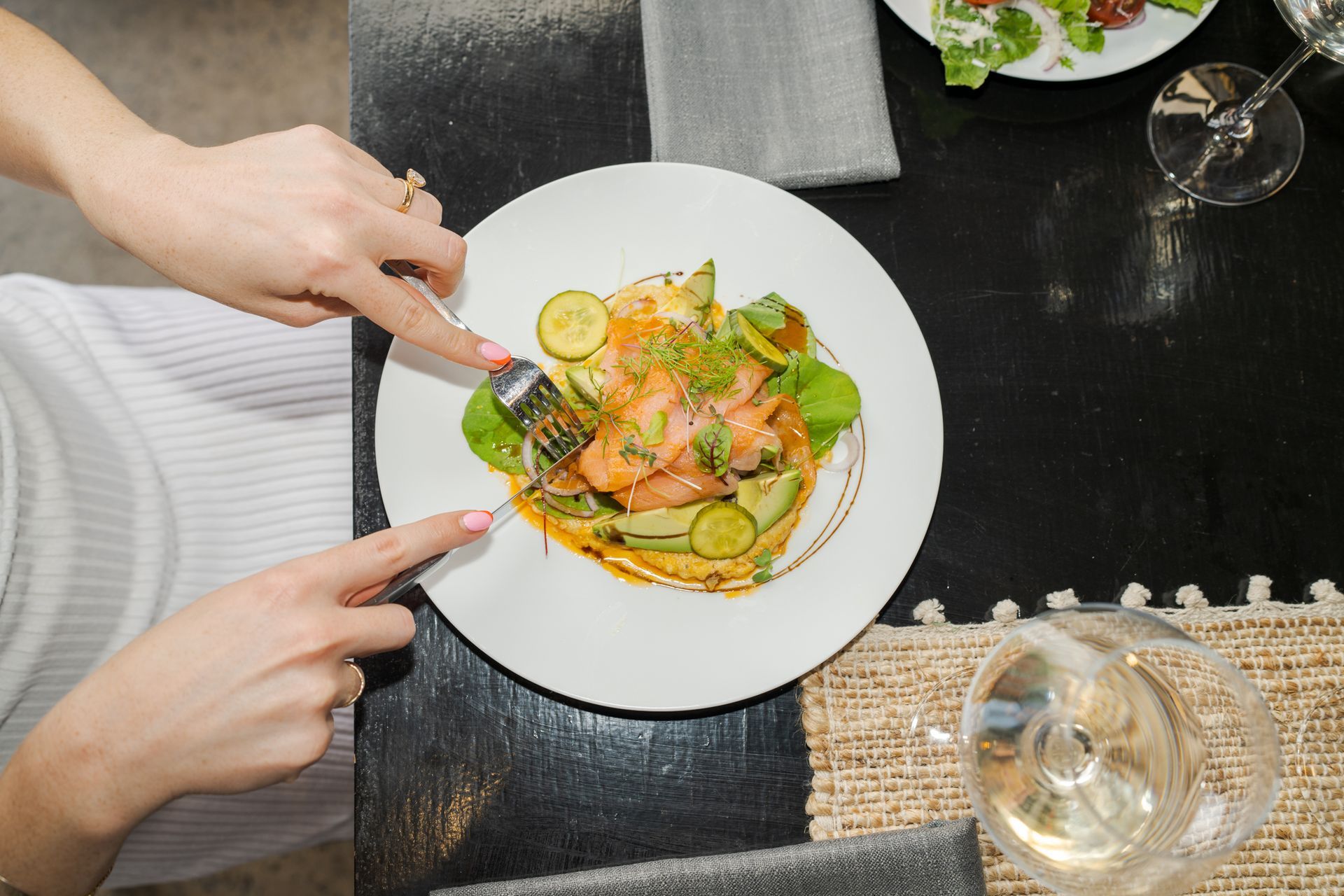 Hands pointing at a plated salmon salad on a restaurant table with wine glasses and a placemat