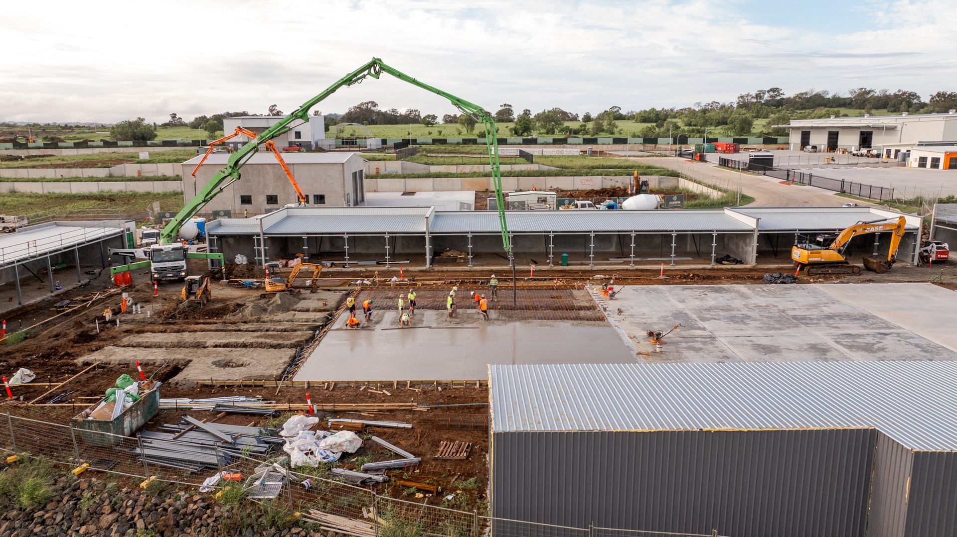 Construction site with workers, cranes, and concrete foundation beside industrial buildings