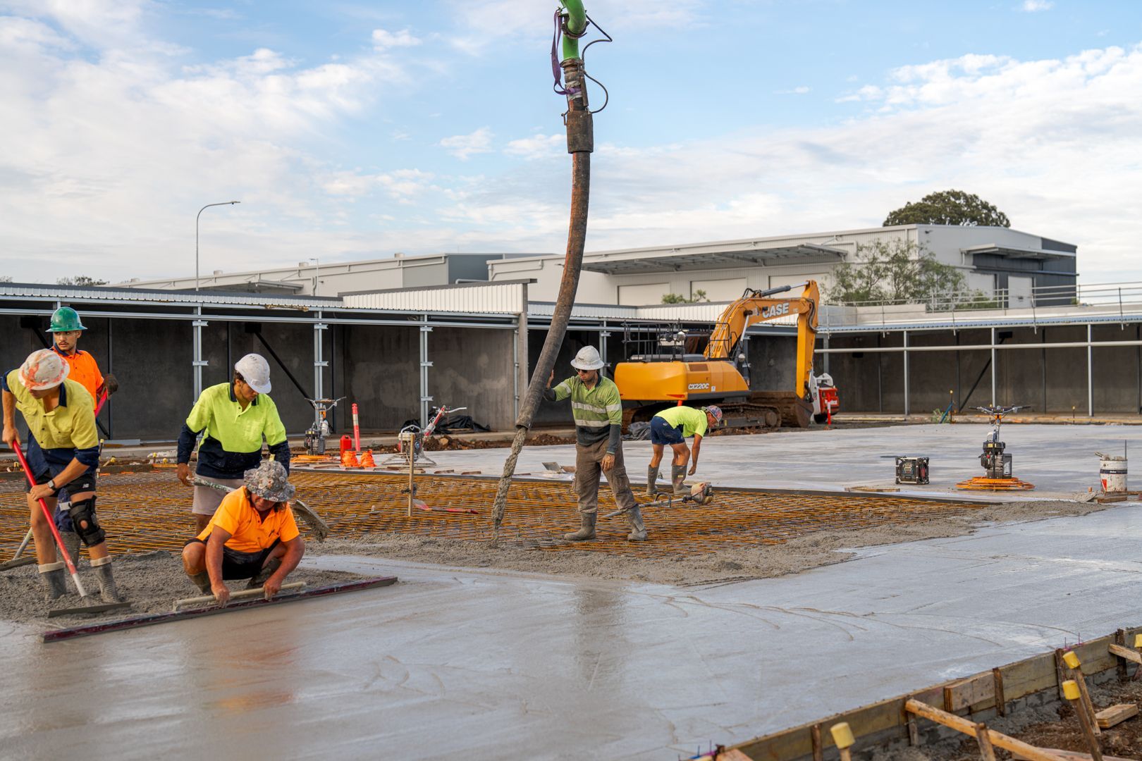 Construction workers smoothing wet concrete on a large slab at an outdoor site with machinery nearby.