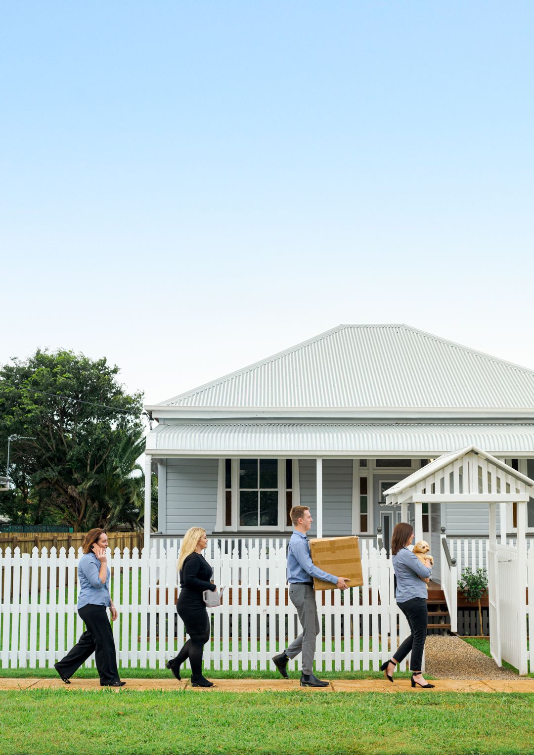 Four people walking past a white house with a picket fence on a sunny day