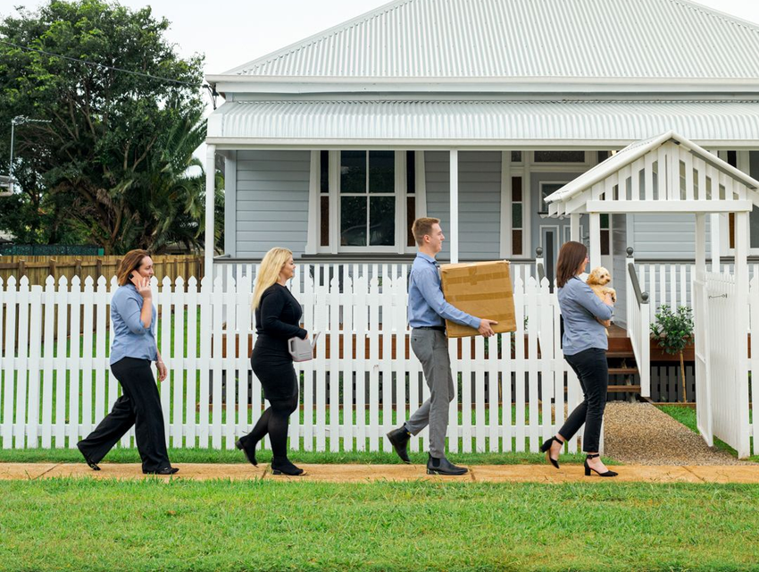 Four people walk past a white picket fence carrying boxes in front of a small house.