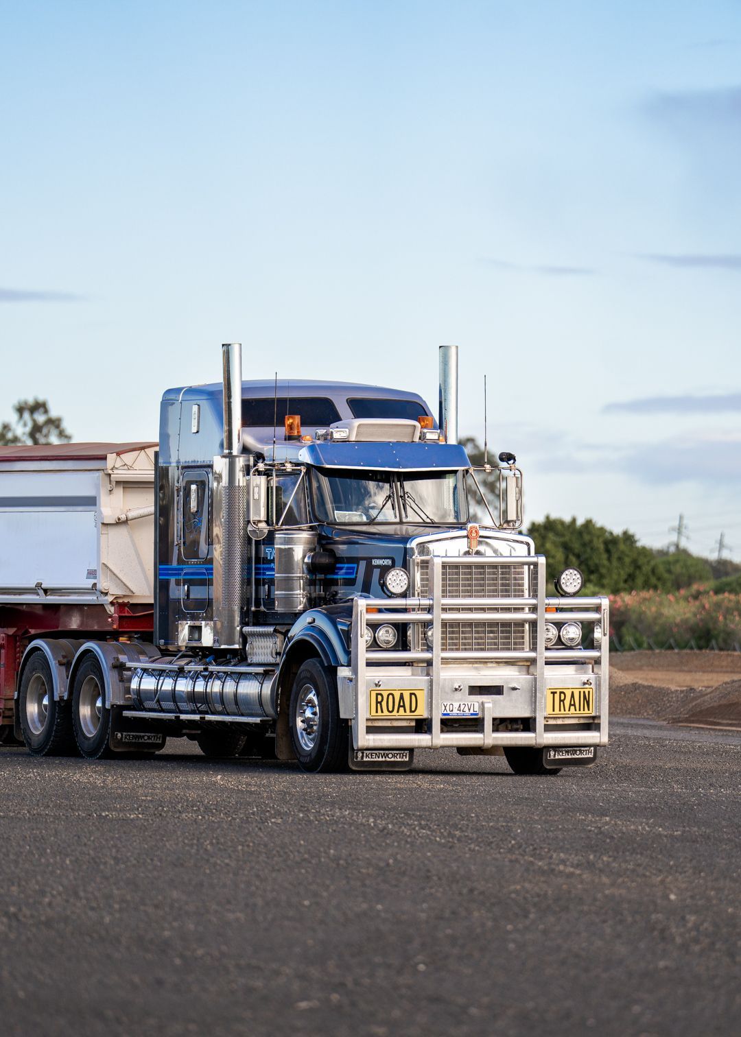 Blue semi-truck with chrome grille and white trailer parked on a gravel lot under a clear sky