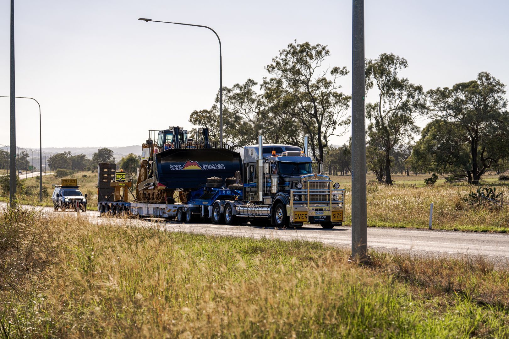 Blue heavy-haul truck towing equipment on a rural road beside grassy fields and trees