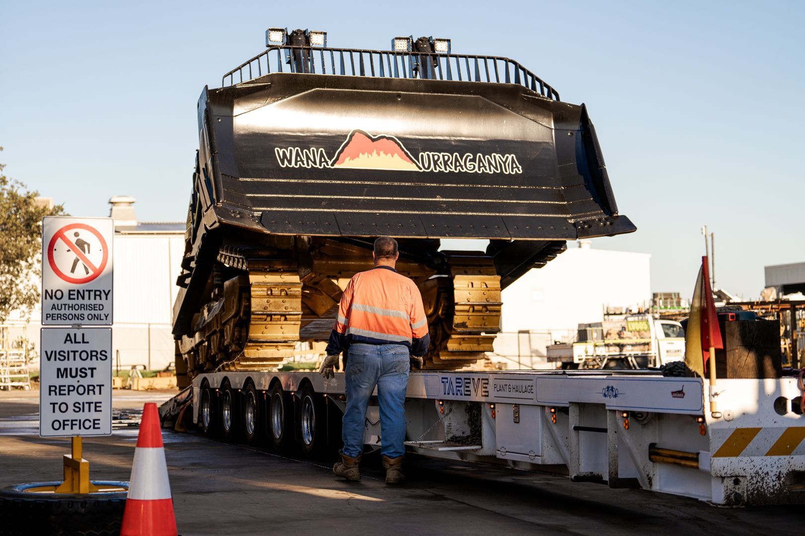 Bulldozer loaded on a flatbed trailer as a worker stands beside it at a roadside worksite