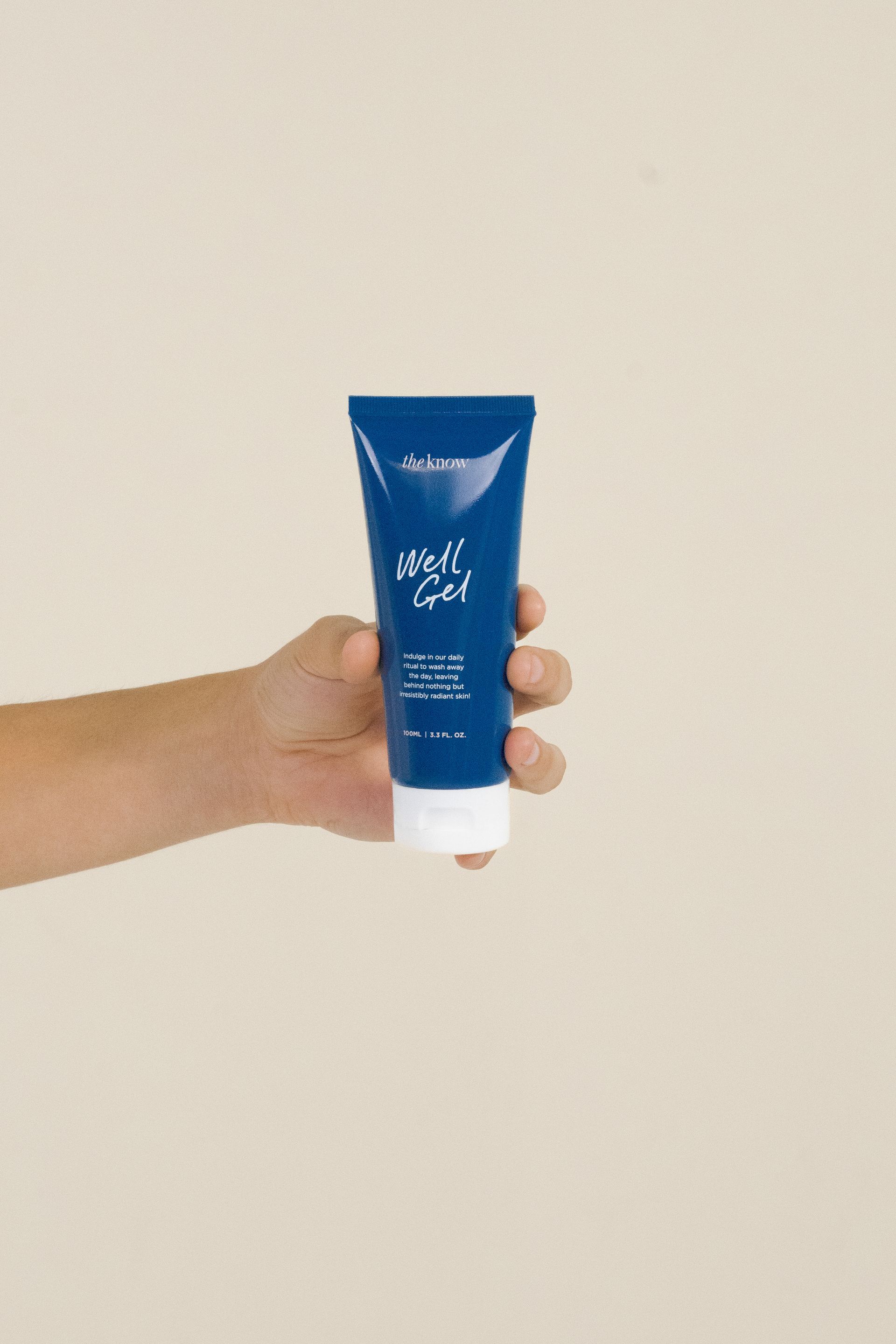 Hand holding a blue tube of cream against a plain beige wall