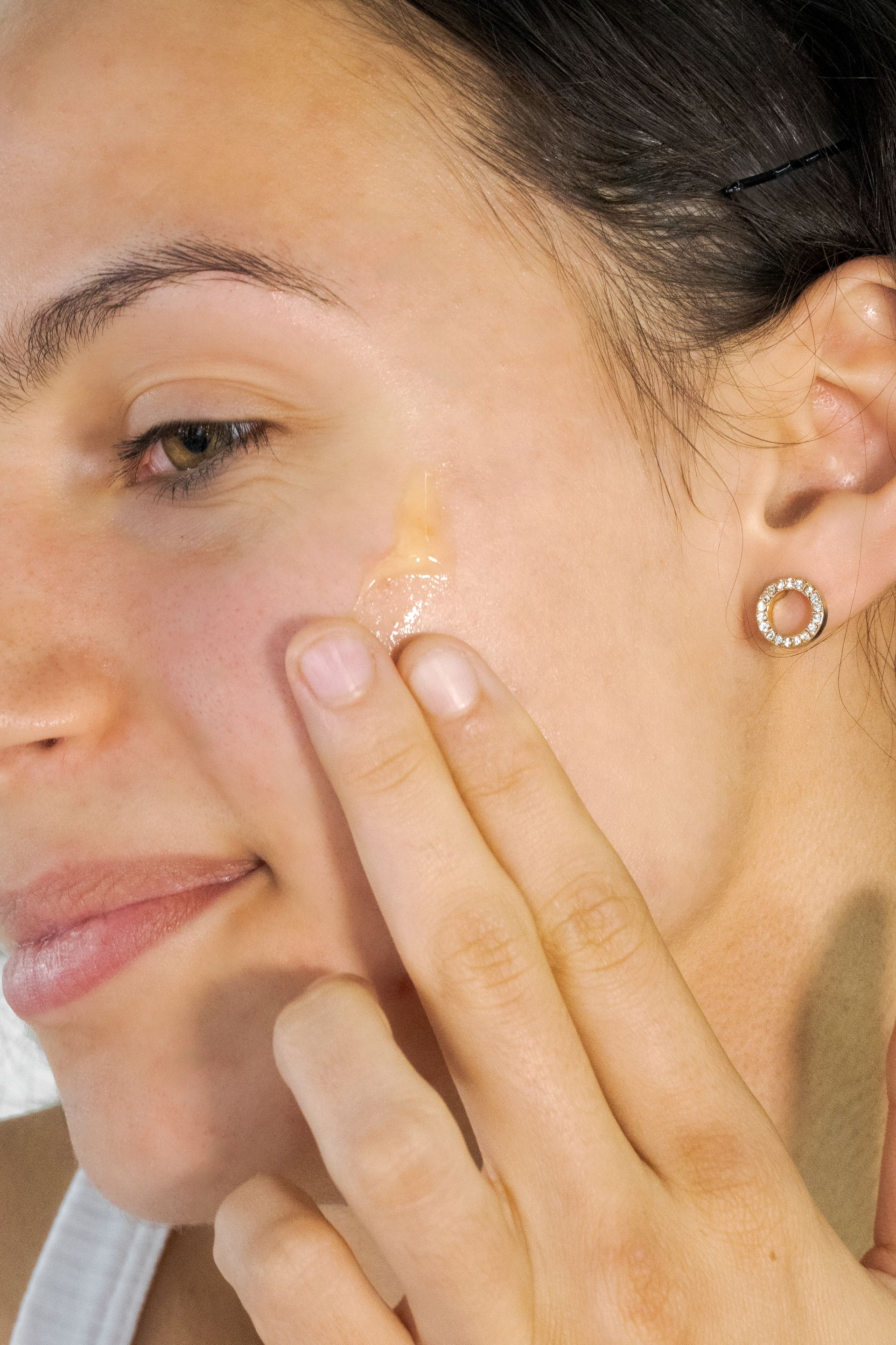 Woman applying cream to her cheek with fingers, wearing a small hoop earring.