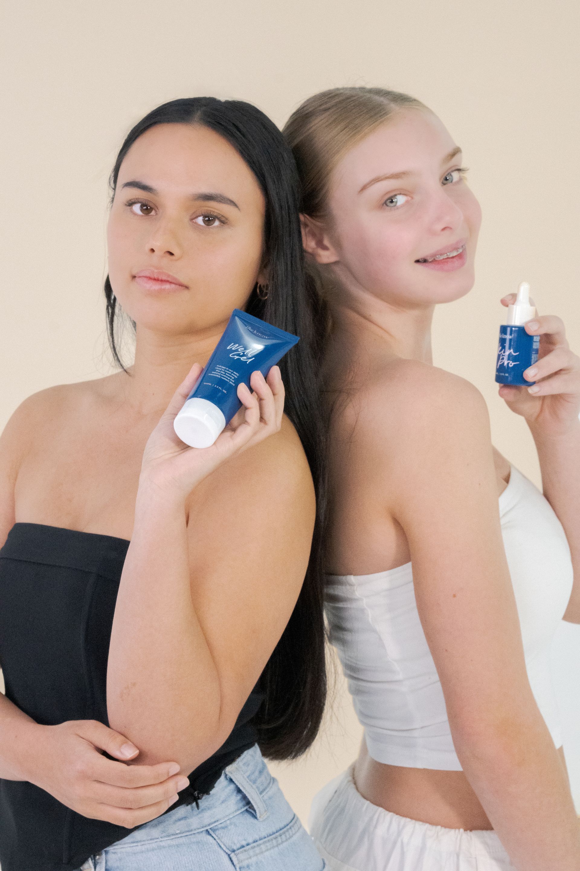 Two models posing back to back, each holding a blue skincare tube on a light background.