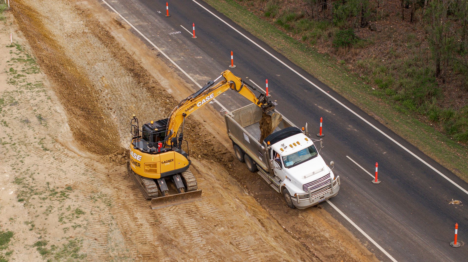 Excavator loading dirt into a white truck beside a road lined with orange cones.