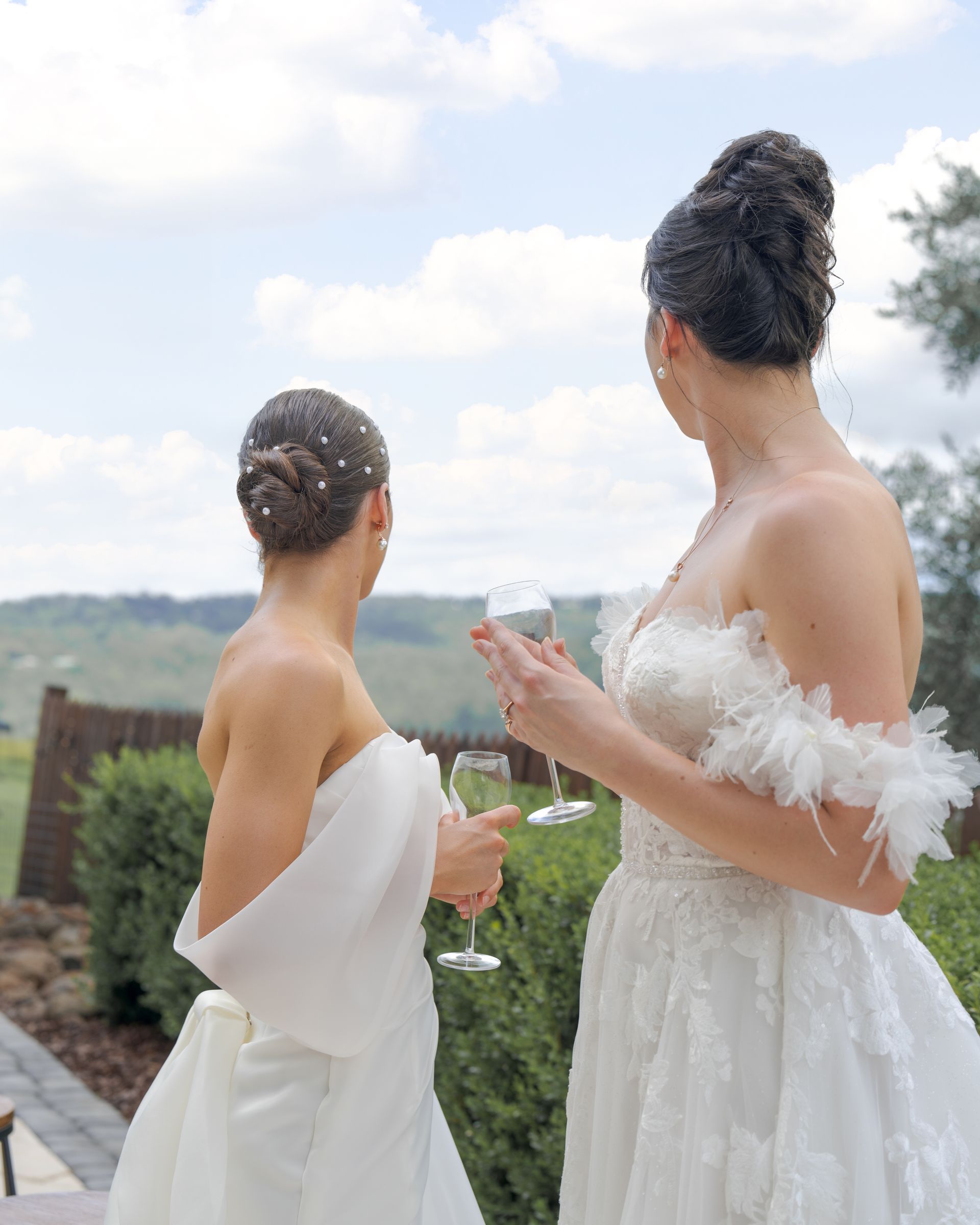 Two brides in white dresses holding drinks, facing a scenic outdoor view at a wedding.