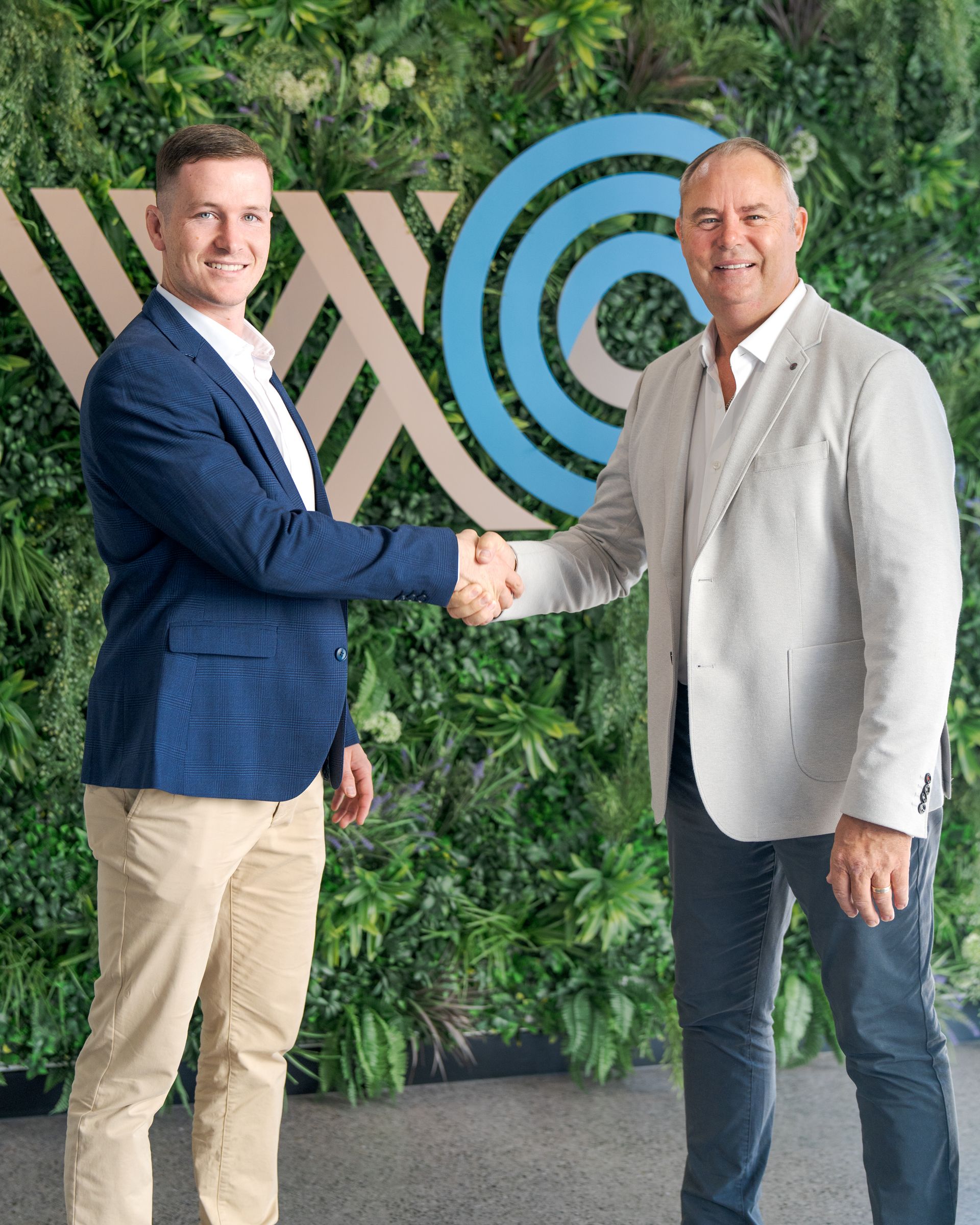 Two men shaking hands in front of a green plant wall and blue circular logo.