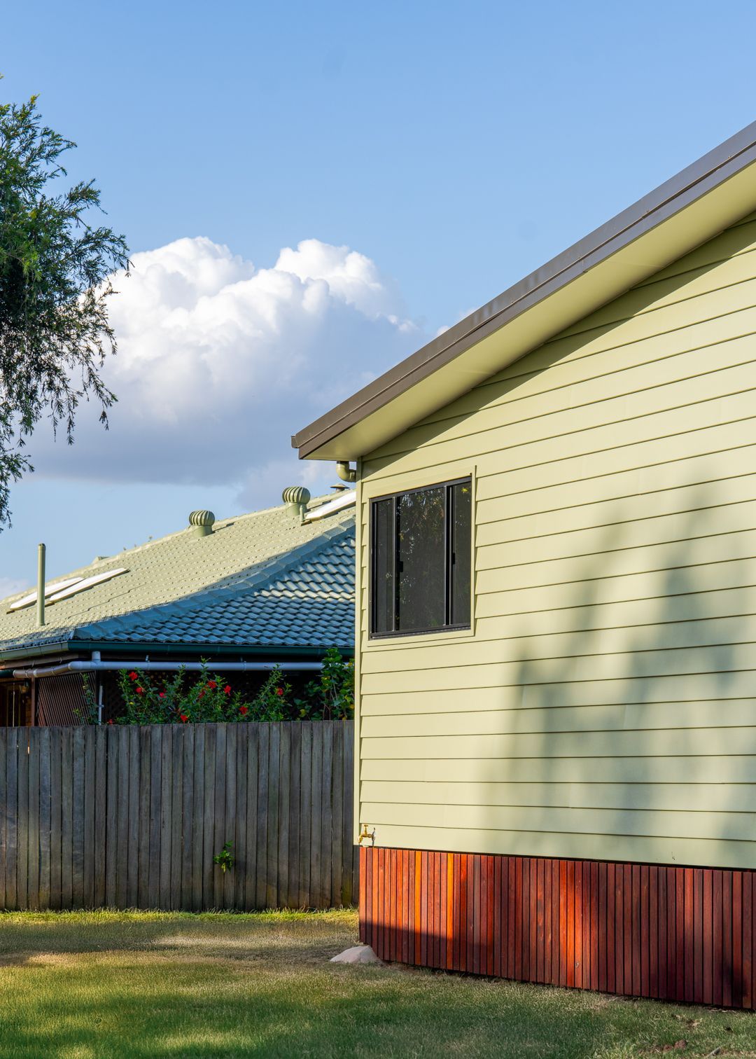 Side of a pale yellow house with red trim and a fenced yard under a blue sky.