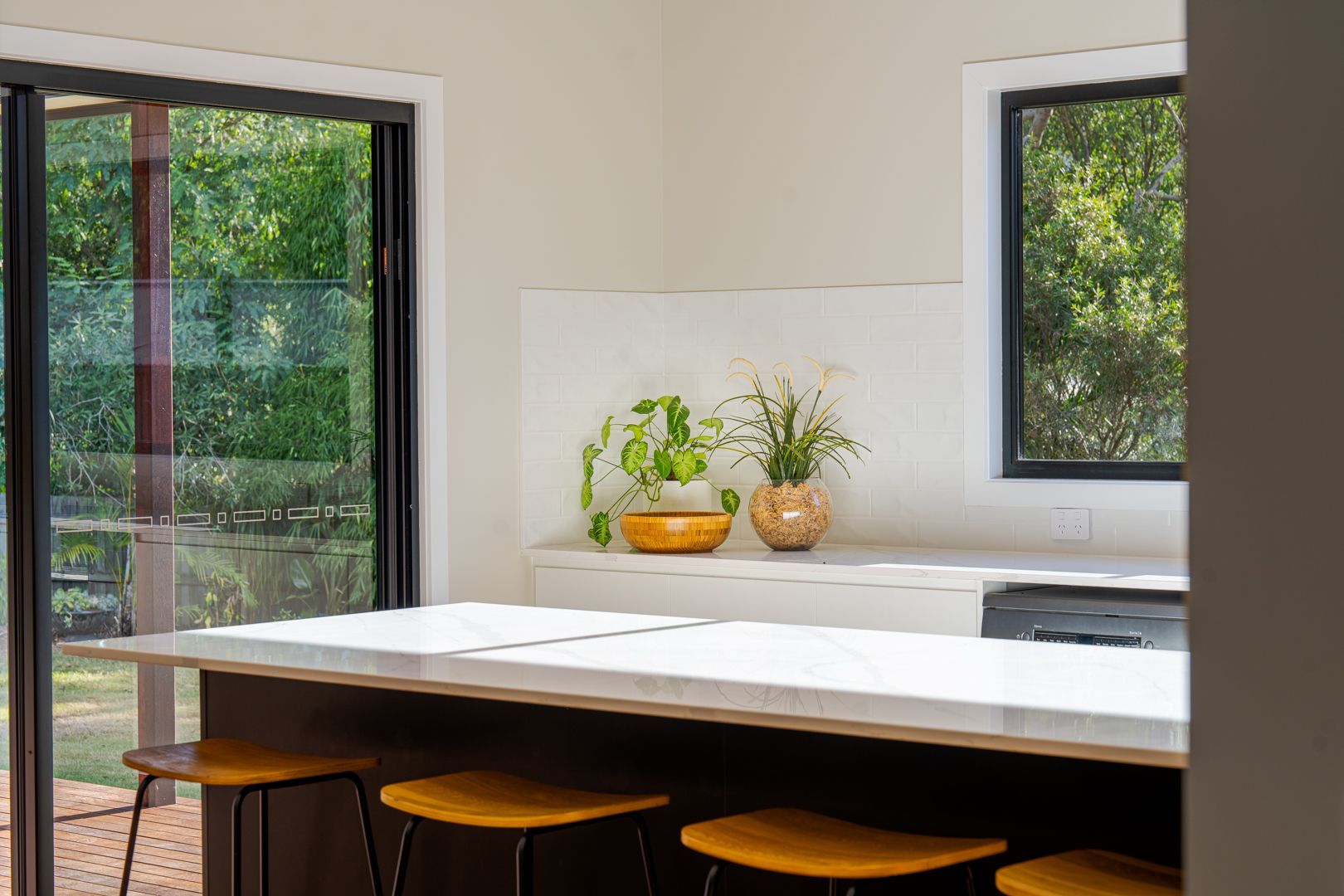 Modern kitchen island with white countertop, black stools, and open sliding doors to a green patio.