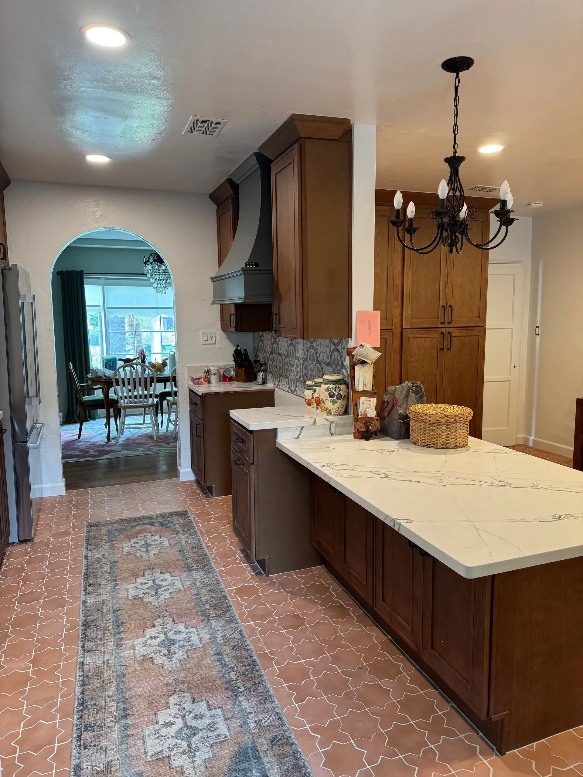 Narrow kitchen with dark wood cabinets, white counters, patterned tile floor, and arched doorway leading to a dining room