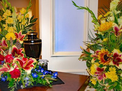 Two colorful floral wreaths beside a black urn on a table, with a white door in the background