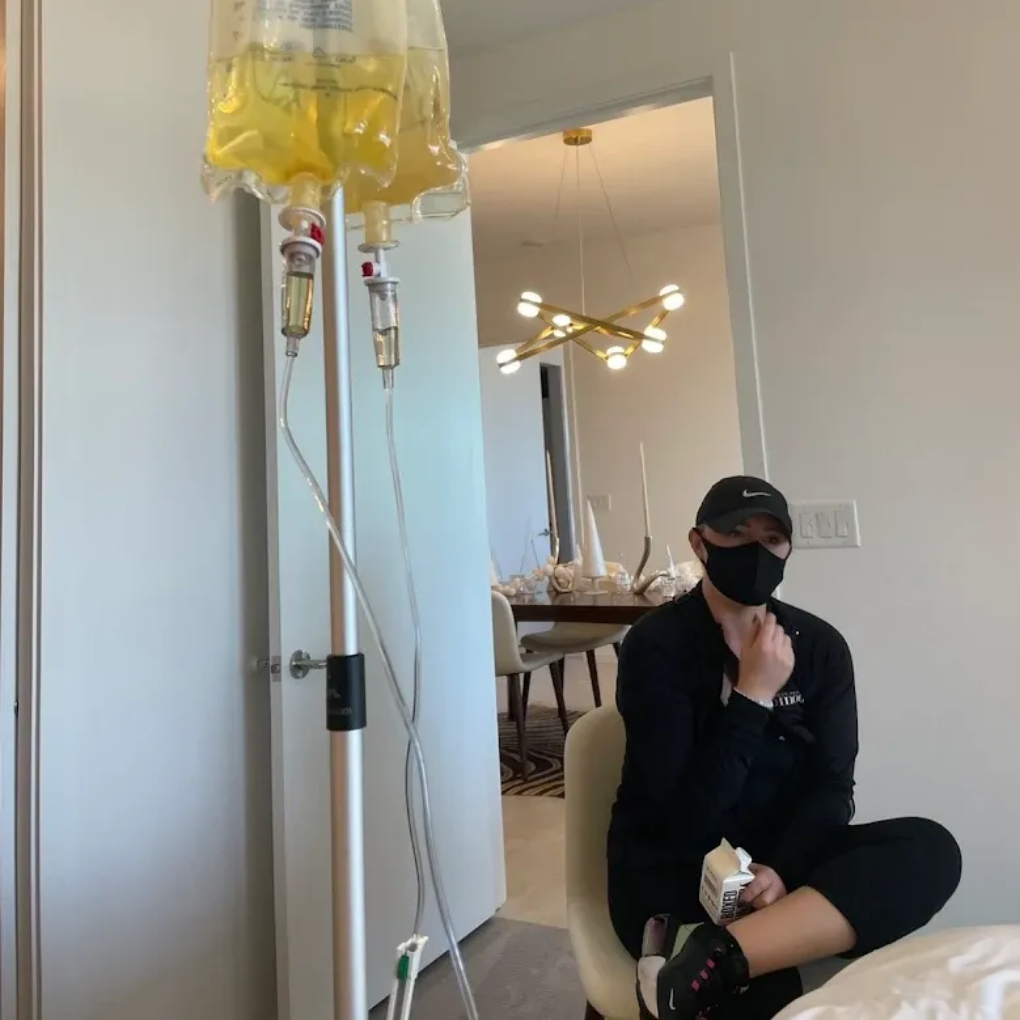 Person wearing mask, receiving IV fluids, sitting near doorway. Bright, modern room with lighting.