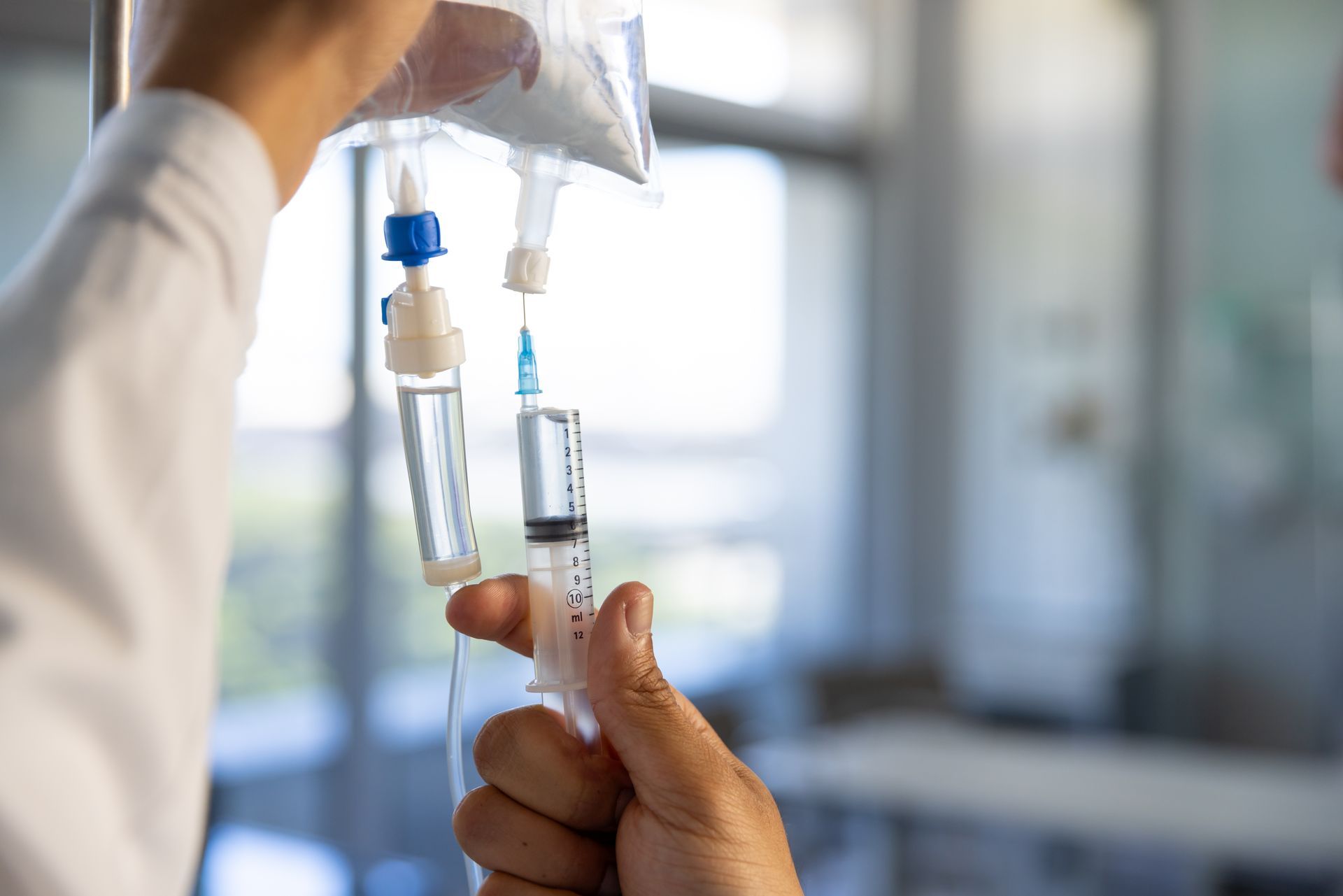Person preparing IV drip bag with a syringe in a bright medical setting.