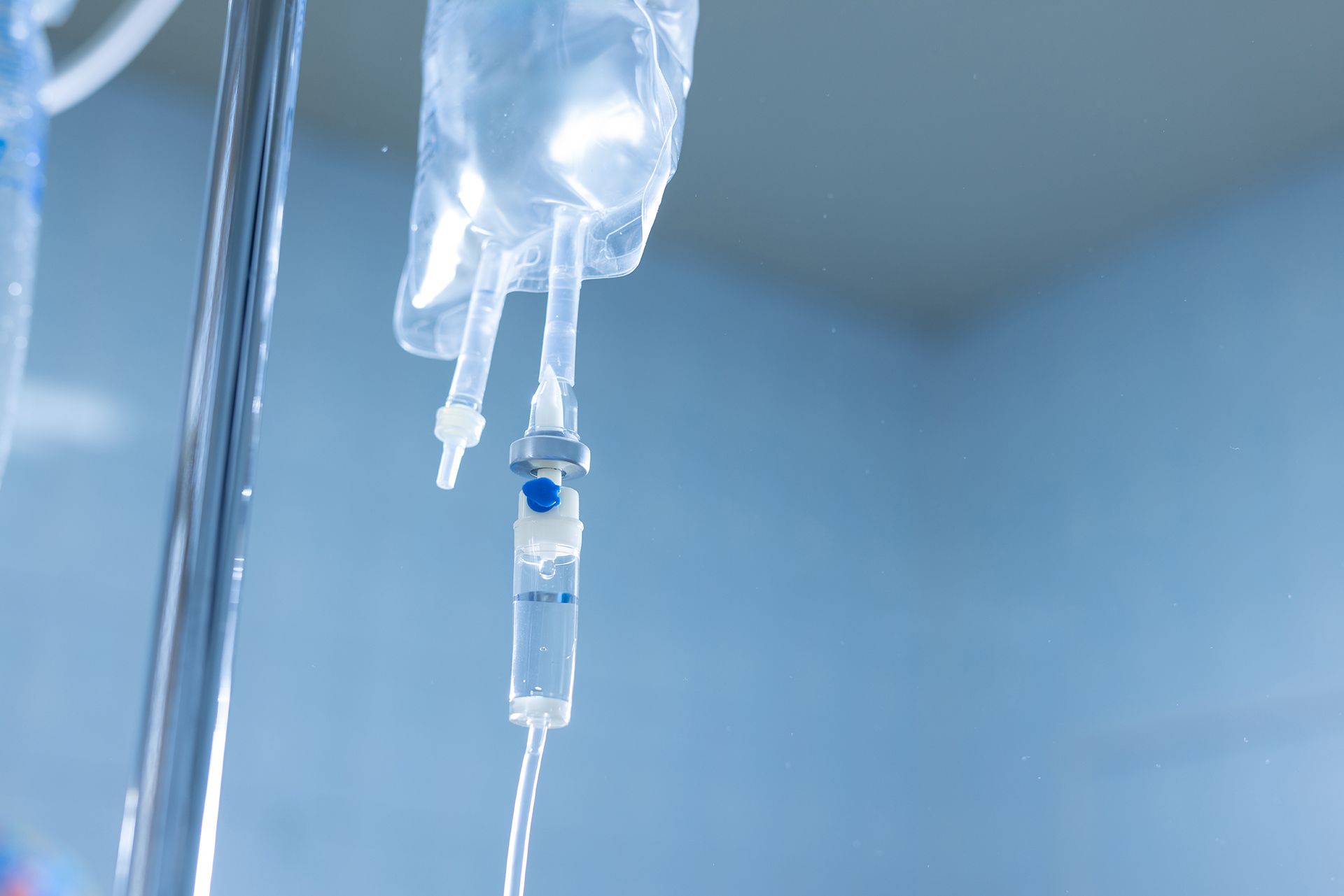 A clear IV bag hangs on a stand, with fluid dripping through a chamber against a blurred, light blue background.