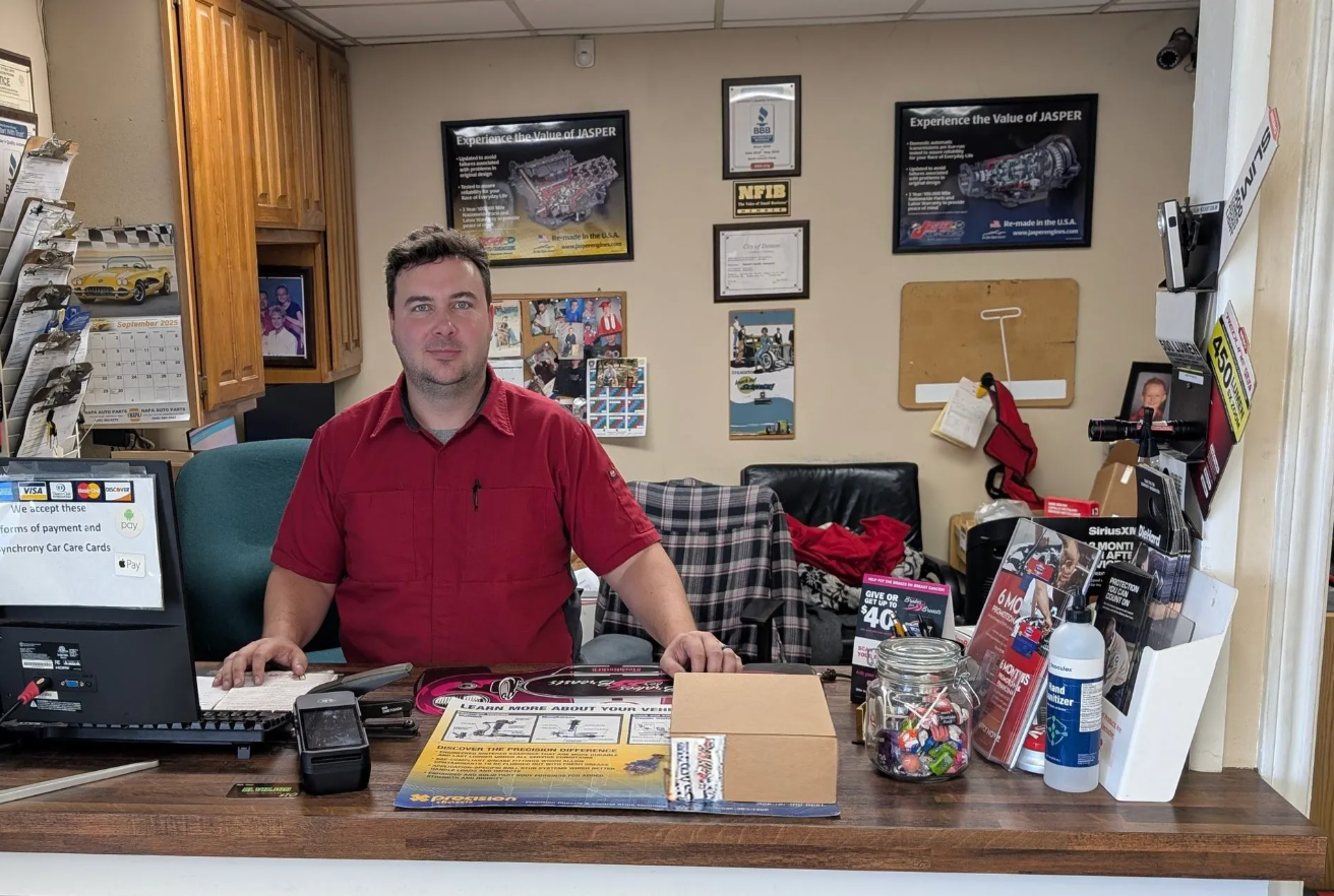 Man behind a shop counter with brochures, mugs, and posters in a small office-like store | Spenser's Quality Automotive