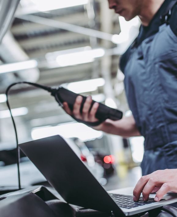 Technician using a handheld scanner while working on a laptop in an industrial setting | OG Autoworks